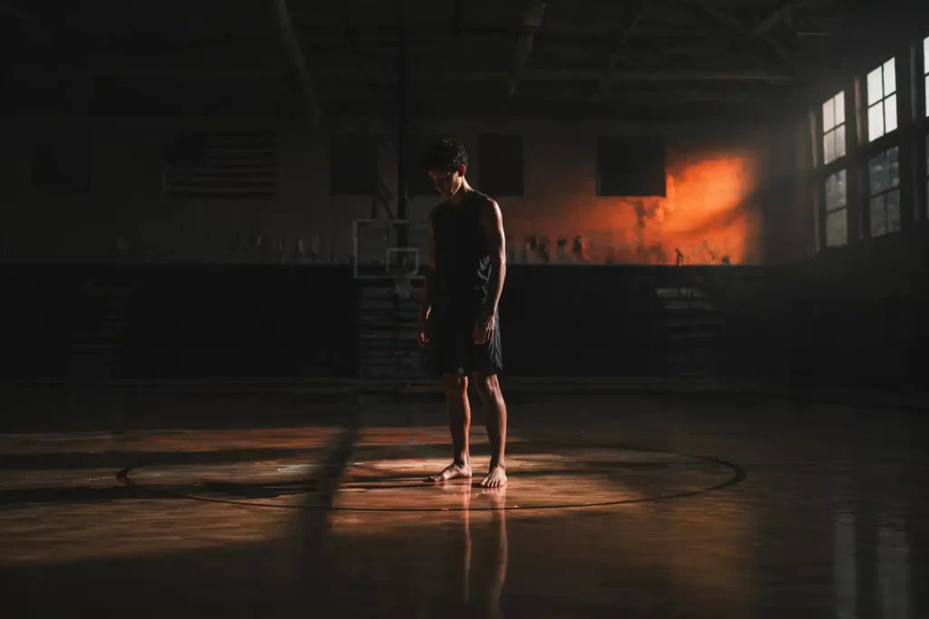 High school basketball player standing barefoot at center court before foot strength training in a dimly lit gymnasium